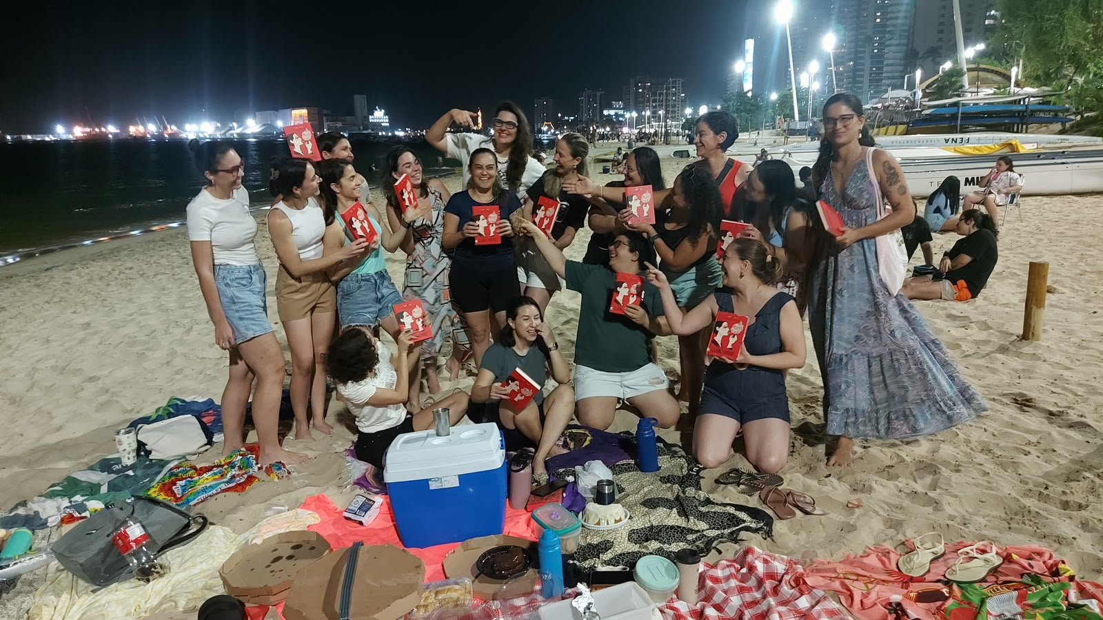 Grupo de leitoras na praia, à noite, segurando exemplares do mesmo livro